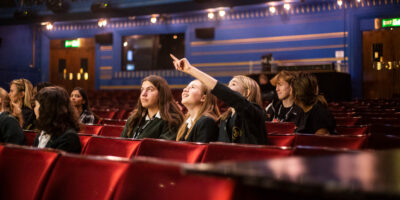 The focus is on three schoolgirls who are sitting in the auditorium of a theatre on red velvet seats. One of the girls is pointing upwards towards something out of the shot and the other two are looking up towards it.