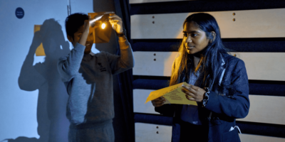 Two young people on stage. One young person is stood holding a light while the second is holding a piece of paper.